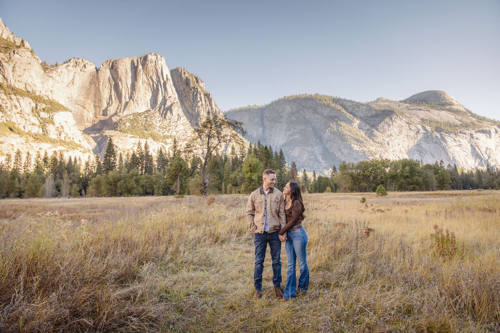 A couple smiles and holds hands in an open field with mountains in the background.