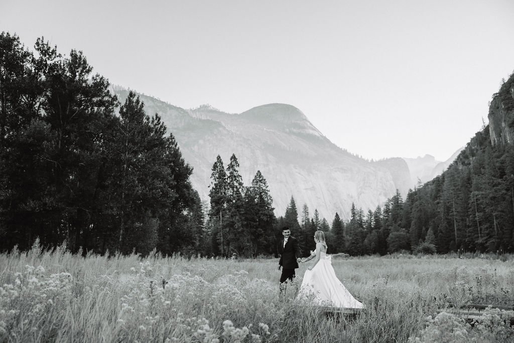 A couple in wedding attire walks through a field with mountains and trees in the background. The image is in black and white.