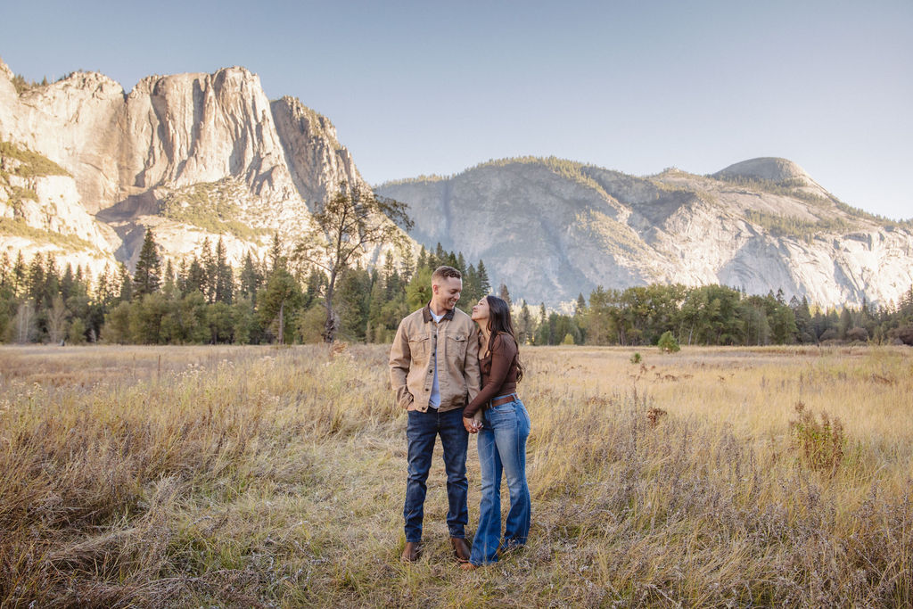 A couple smiles and holds hands in an open field with mountains in the background.