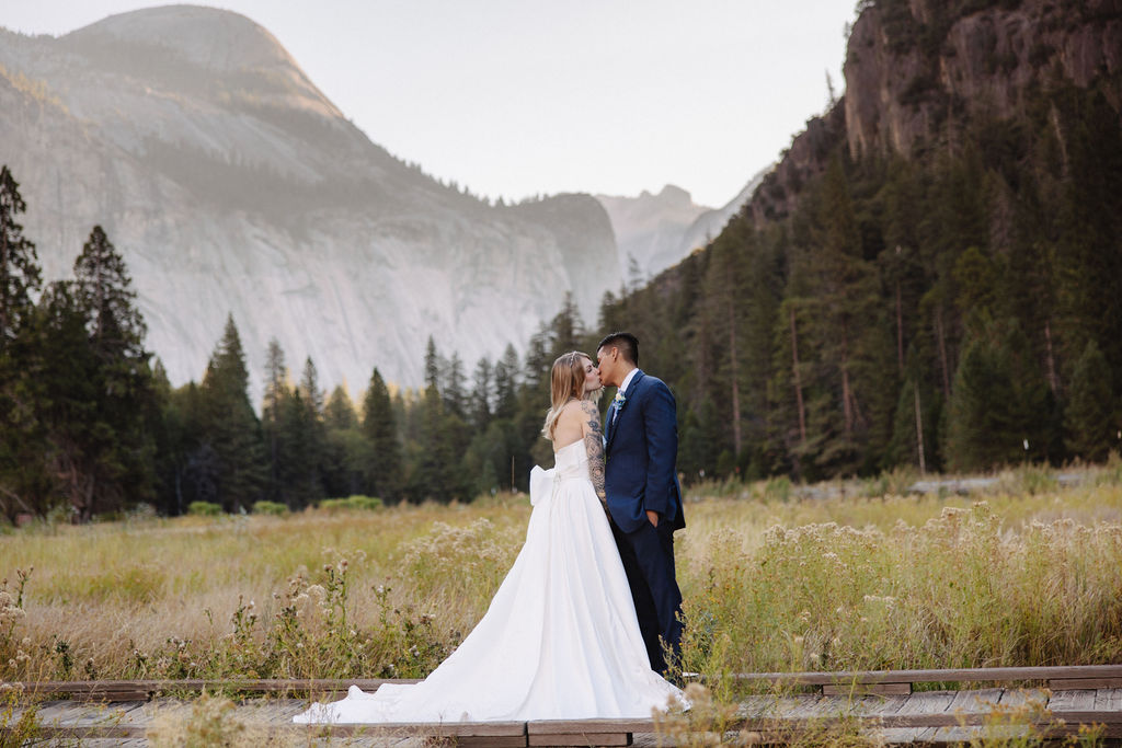 A couple in wedding attire walks through a field with mountains and trees in the background. 