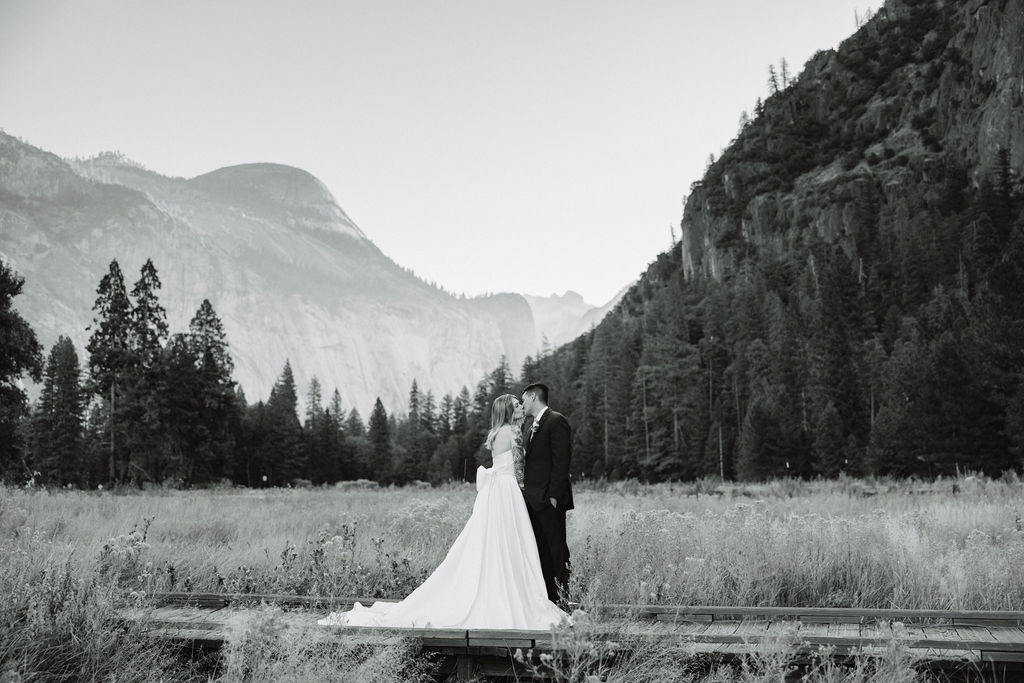 A couple in wedding attire walks through a field with mountains and trees in the background for an elopement at bridalveil falls