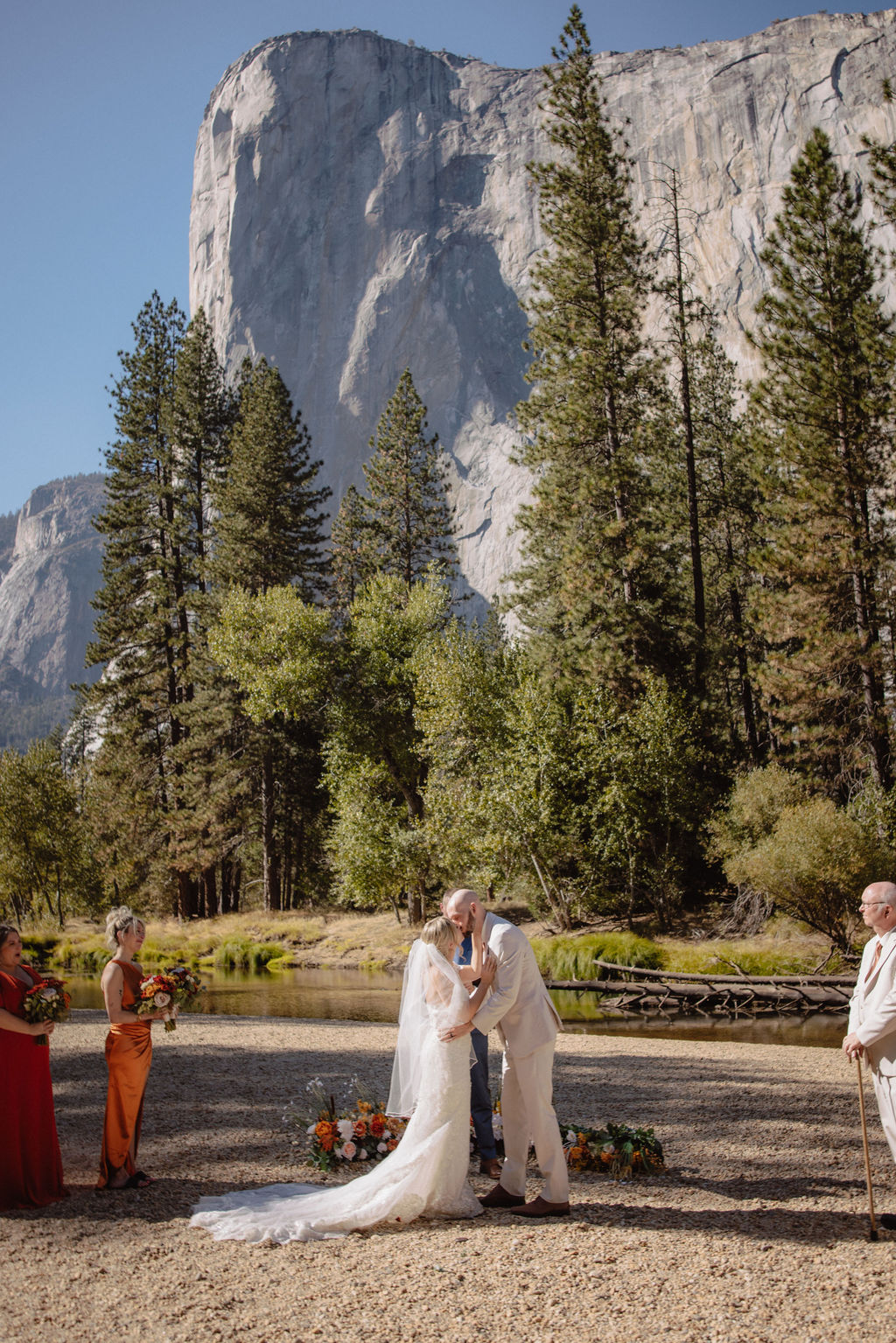 A couple exchanges vows outdoors surrounded by guests, with tall pine trees and a large mountain in the background.