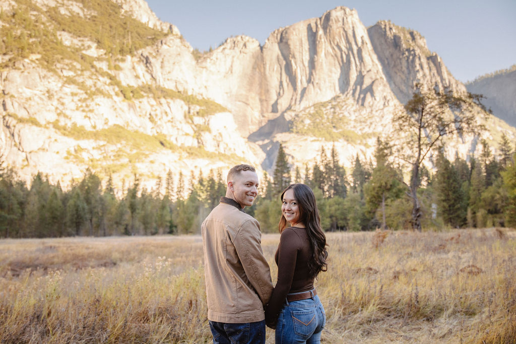 A couple smiles and holds hands in an open field with mountains in the background.