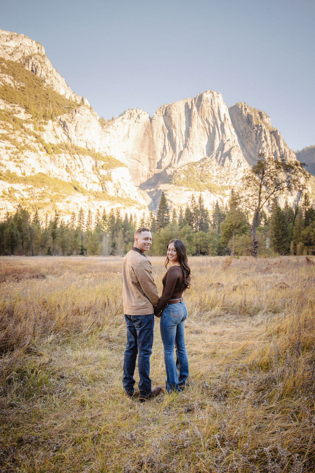 A couple smiles and holds hands in an open field with mountains in the background.