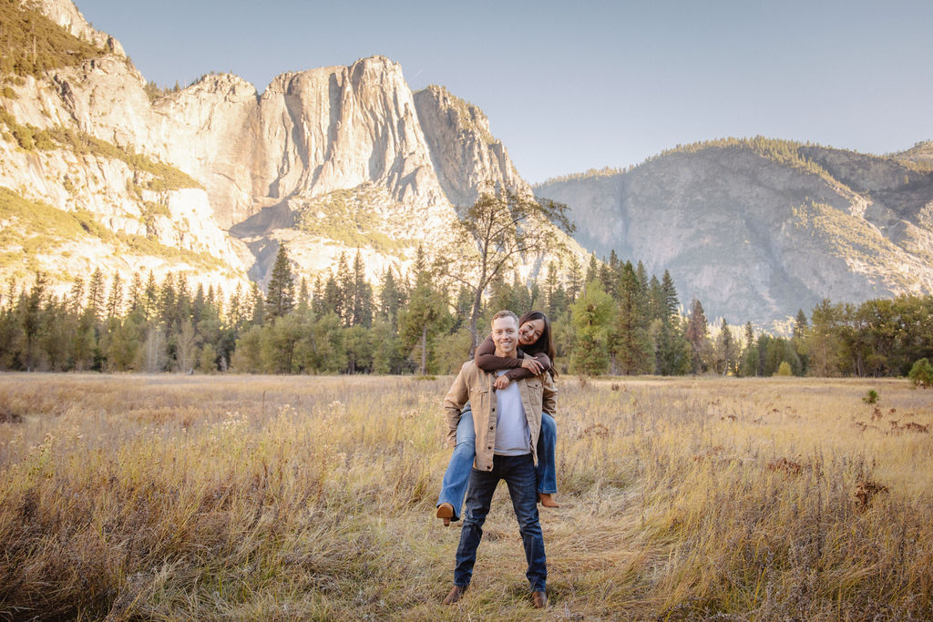 A couple smiles and holds hands in an open field with mountains in the background.