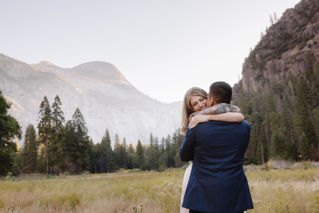 A couple in wedding attire walks through a field with mountains and trees in the background for an elopement at bridalveil falls