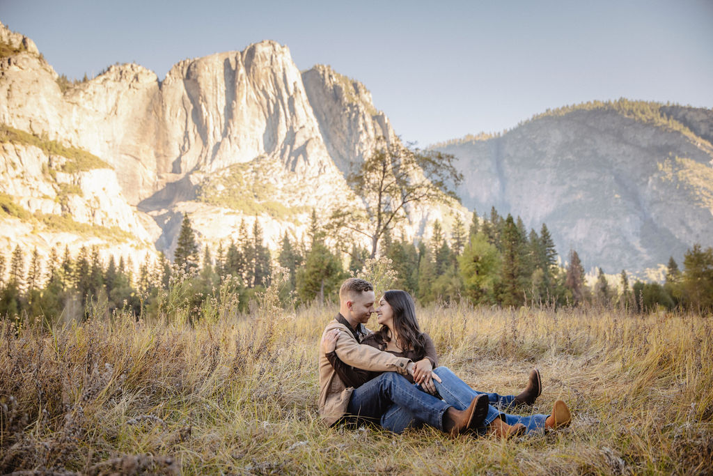 A couple smiles and holds hands in an open field with mountains in the background.