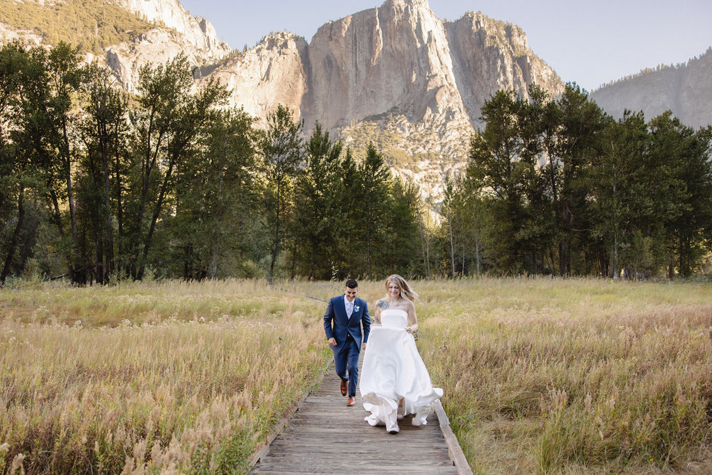 A couple in wedding attire walks through a field with mountains and trees in the background for an elopement at bridalveil falls