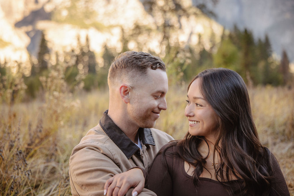 A couple smiles and holds hands in an open field with mountains in the background.