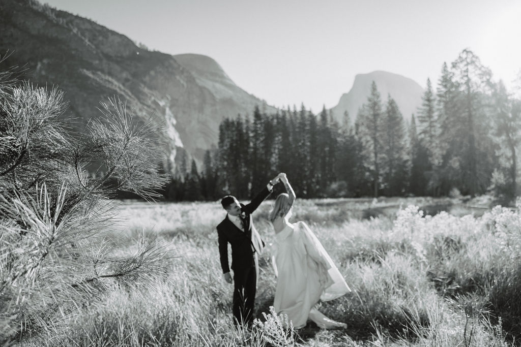 A couple in wedding attire walks through a field with mountains and trees in the background for an elopement at bridalveil falls