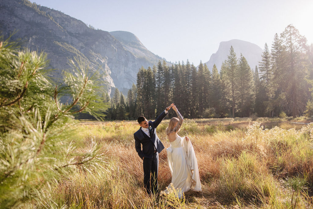 A couple in wedding attire walks through a field with mountains and trees in the background for an elopement at bridalveil falls