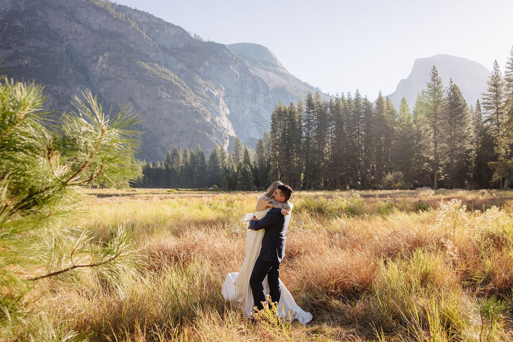  A couple in wedding attire walks through a field with mountains and trees in the background for an elopement at bridalveil falls