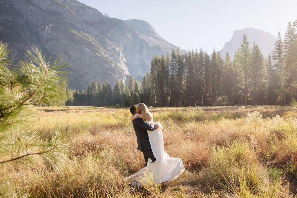 A couple in wedding attire walks through a field with mountains and trees in the background for an elopement at bridalveil falls