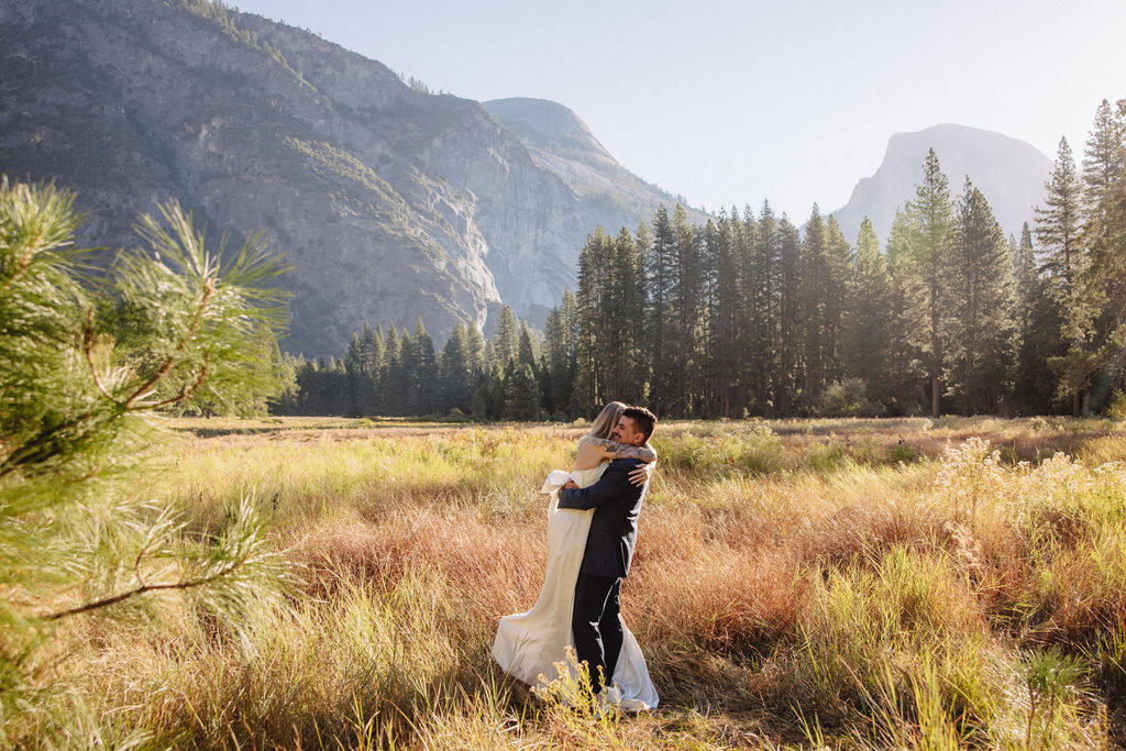 A couple in wedding attire walks through a field with mountains and trees in the backgroundfor an elopement at bridalveil falls