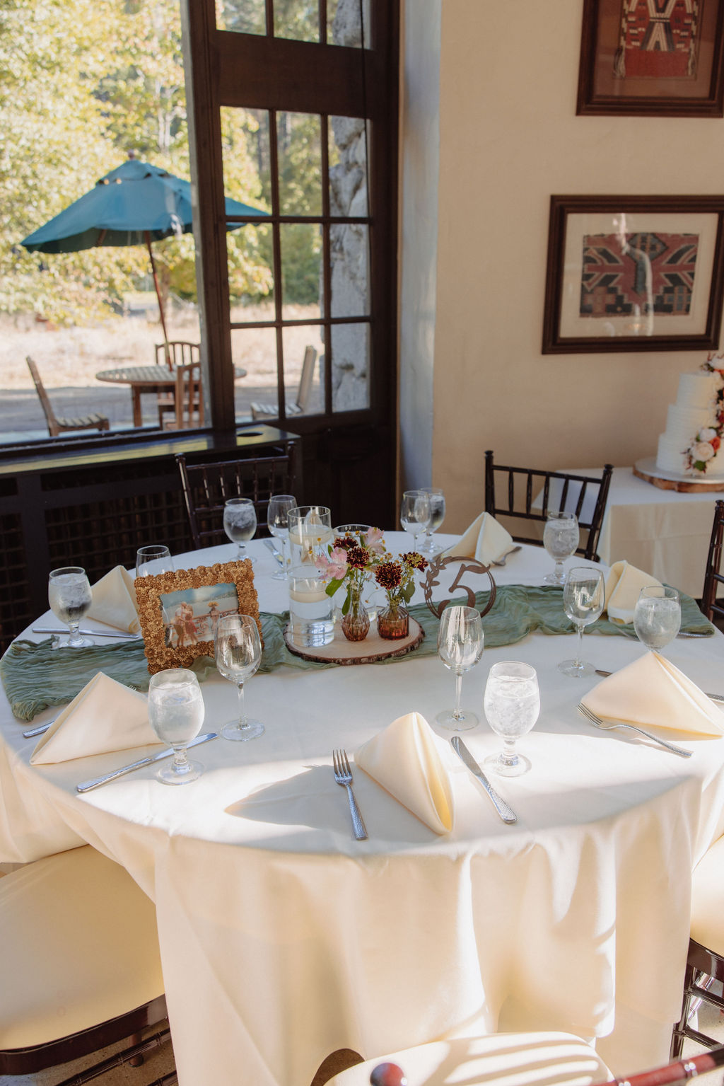 A round table set for a formal event with white tablecloth, napkins, glassware, and a centerpiece. There are paintings on the wall and a view of a patio with a green umbrella outside.