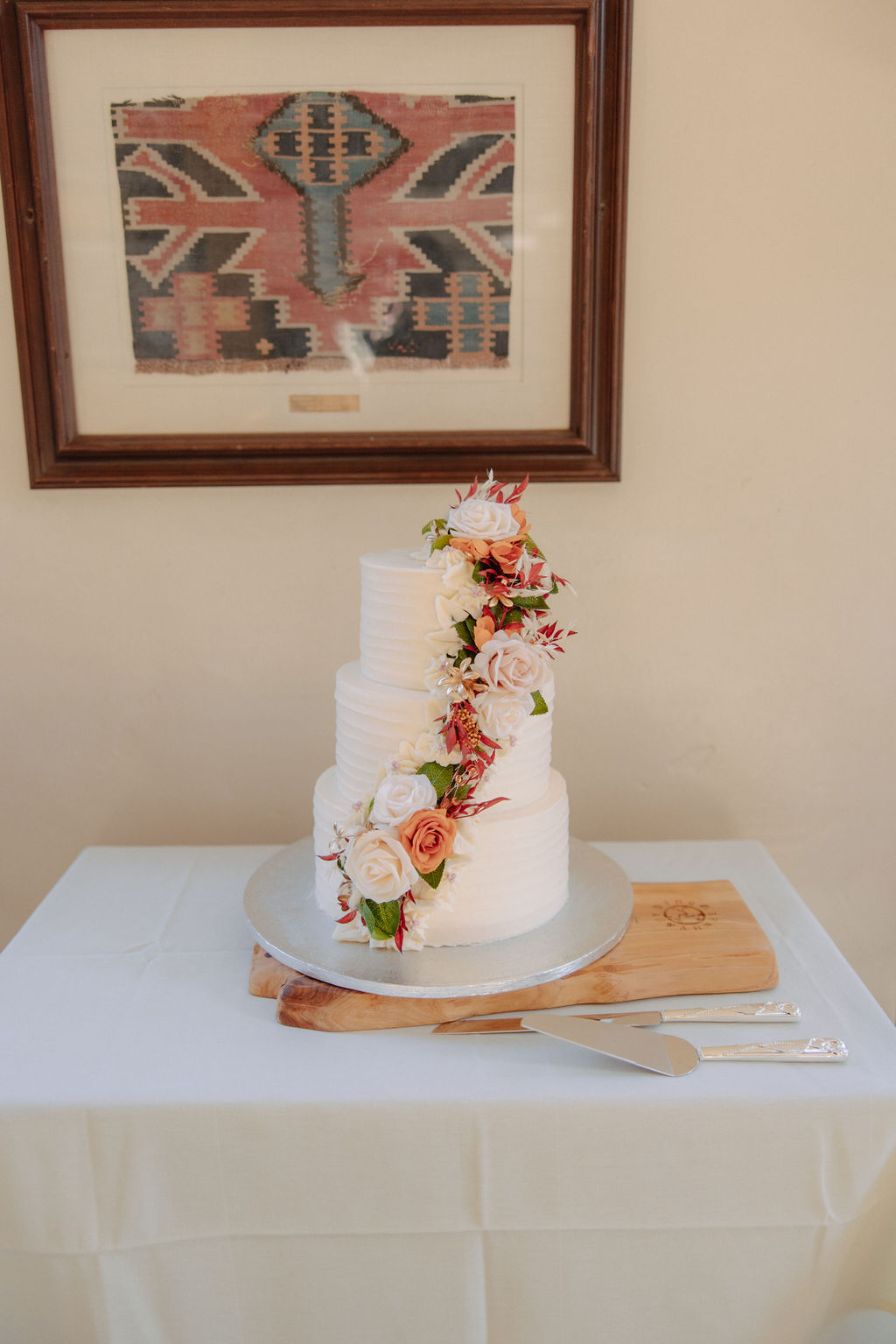 A three-tier white cake with floral decoration is placed on a table with a cake knife. A framed artwork hangs on the wall behind it.