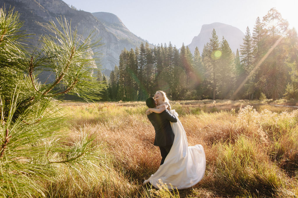 A couple in wedding attire walks through a field with mountains and trees in the background for an elopement at bridalveil falls