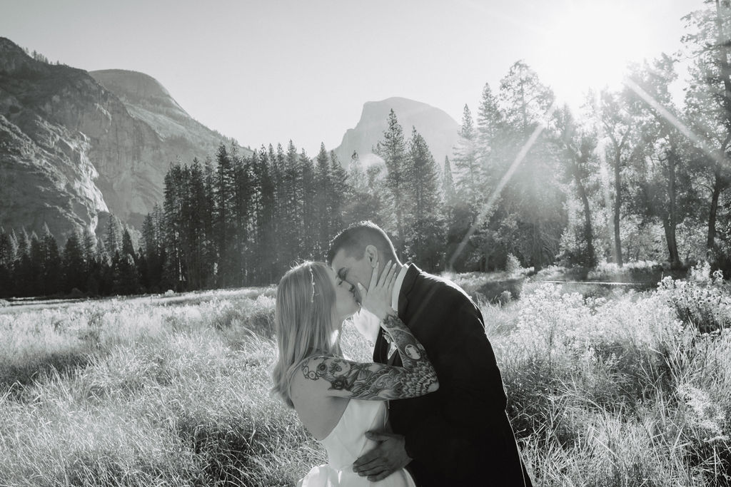 A couple in wedding attire walks through a field with mountains and trees in the background for an elopement at bridalveil falls