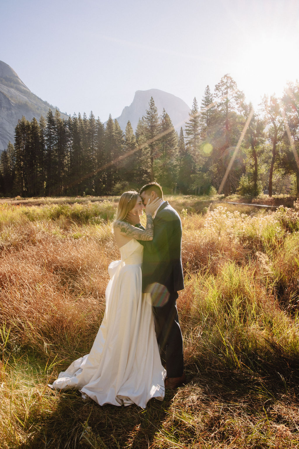 A couple in wedding attire walks through a field with mountains and trees in the background for an elopement at bridalveil falls