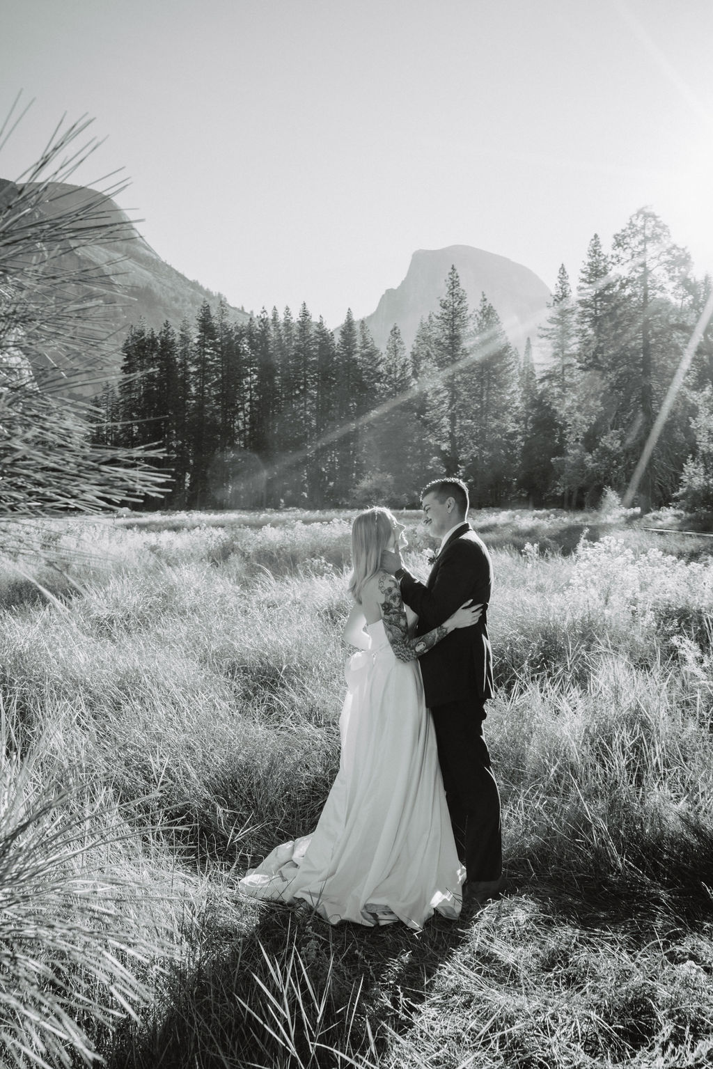 A couple in wedding attire walks through a field with mountains and trees in the background for an elopement at bridalveil falls