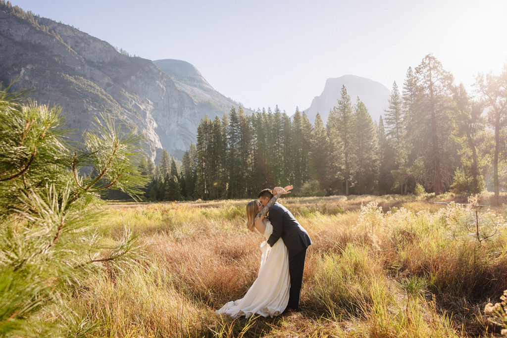 A couple in wedding attire walks through a field with mountains and trees in the background for an elopement at bridalveil falls