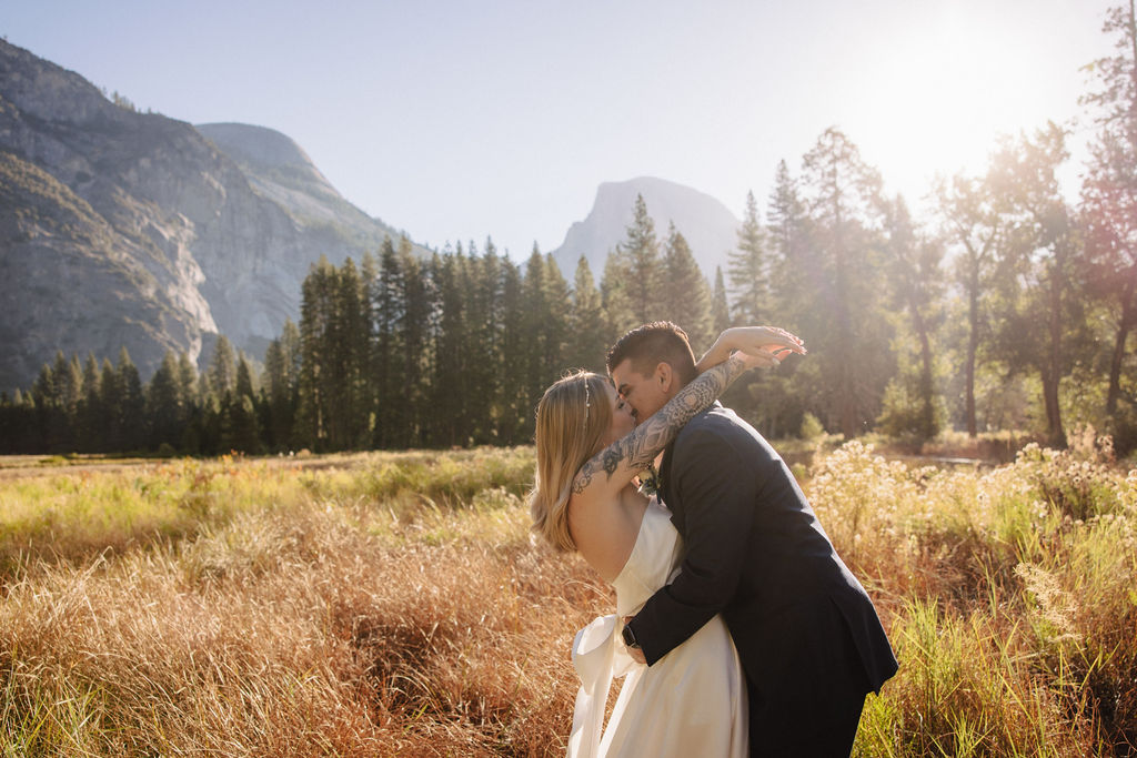 A couple in wedding attire walks through a field with mountains and trees in the background for an elopement at bridalveil falls