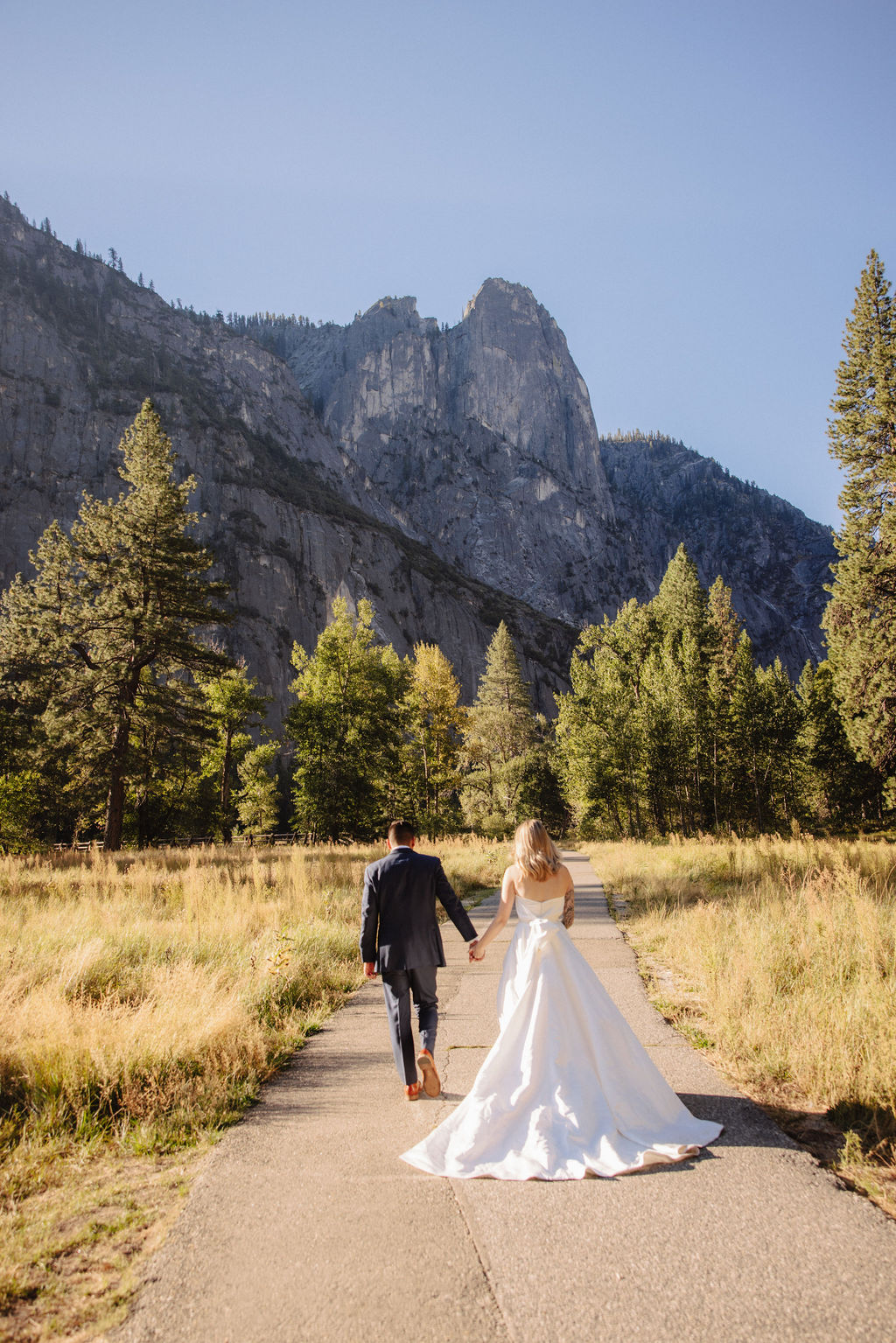 A couple in wedding attire walks through a field with mountains and trees in the background for an elopement at bridalveil falls