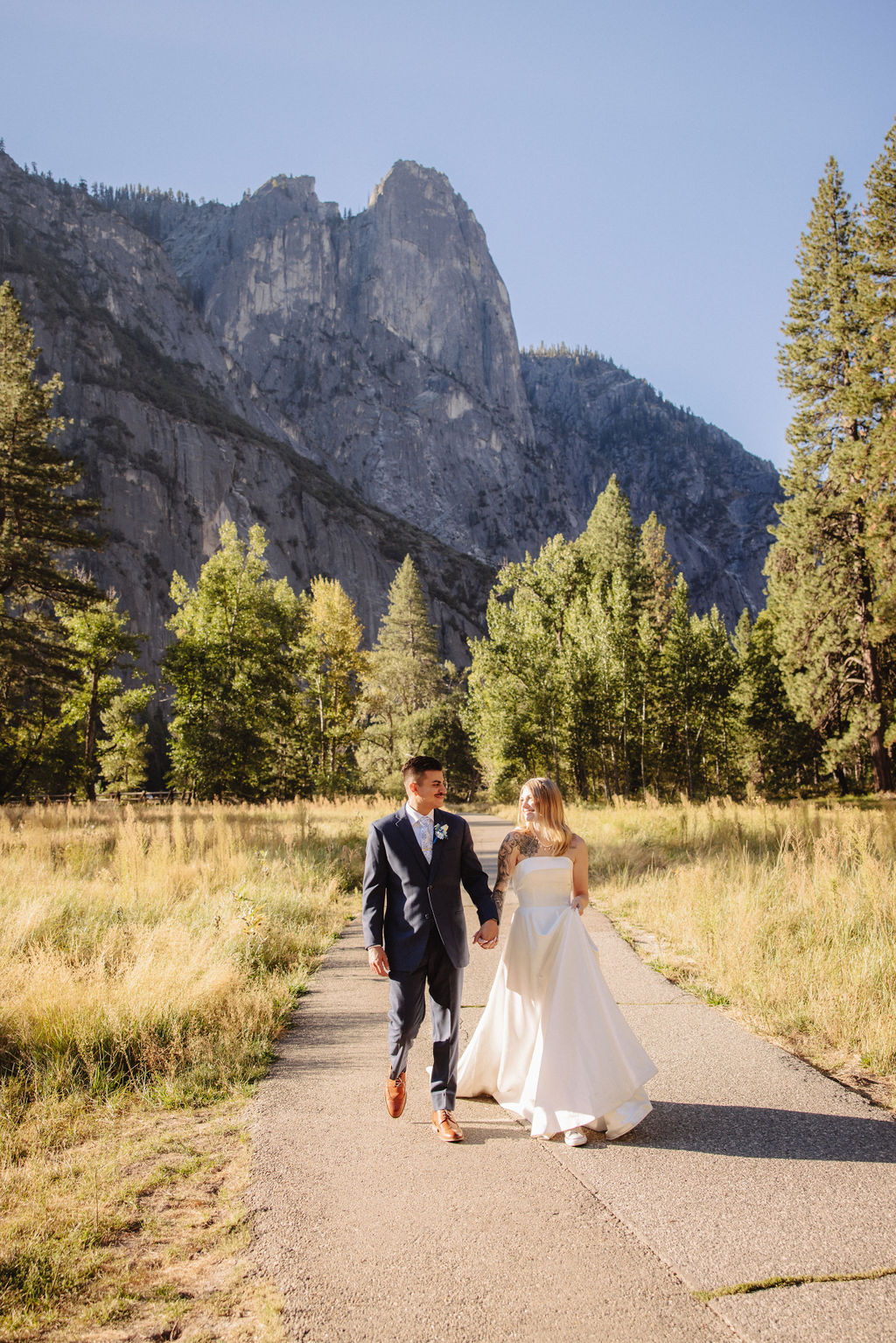 A couple in wedding attire walks through a field with mountains and trees in the background for an elopement at bridalveil falls