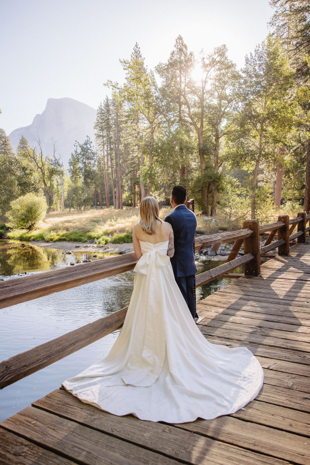 A couple in wedding attire walks through a field with mountains and trees in the background for an elopement at bridalveil falls