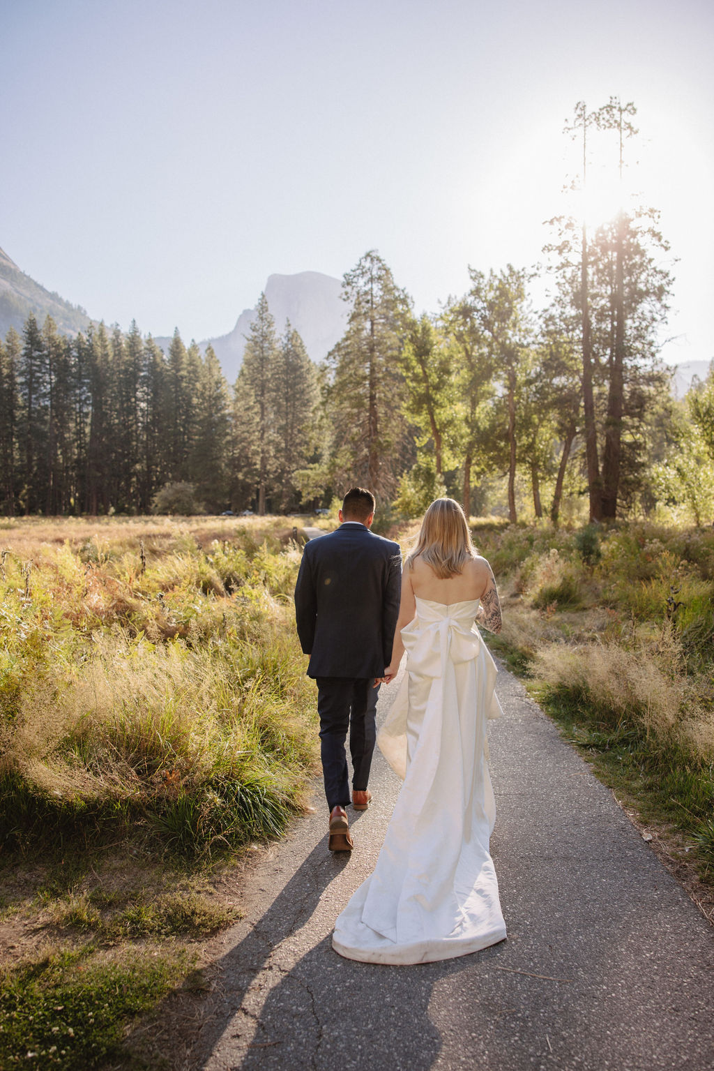 A couple in wedding attire walks through a field with mountains and trees in the background for an elopement at bridalveil falls