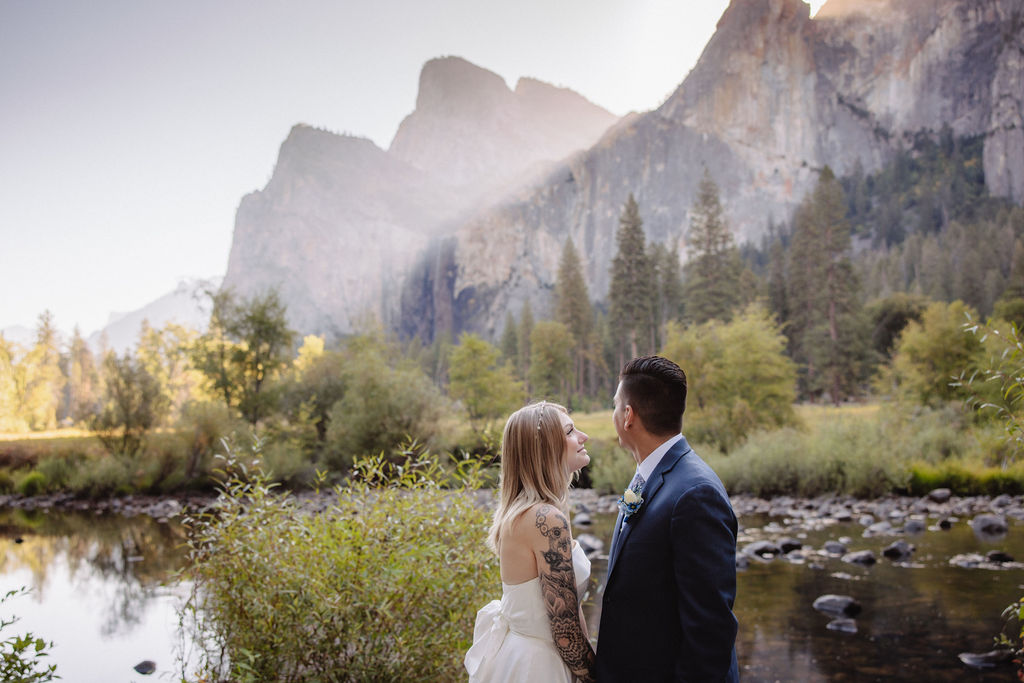 A couple in wedding attire walks through a field with mountains and trees in the background for an elopement at bridalveil falls