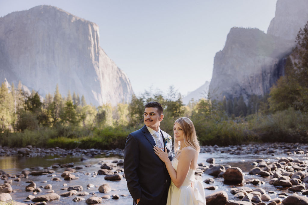 A couple in wedding attire walks through a field with mountains and trees in the background for an elopement at bridalveil falls