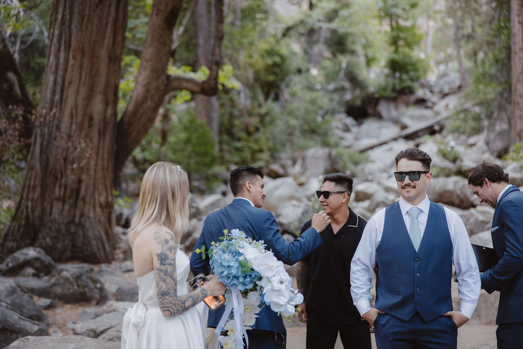 A bride holds a bouquet while four men in suits and sunglasses stand and talk in a wooded area with rocks in the background.