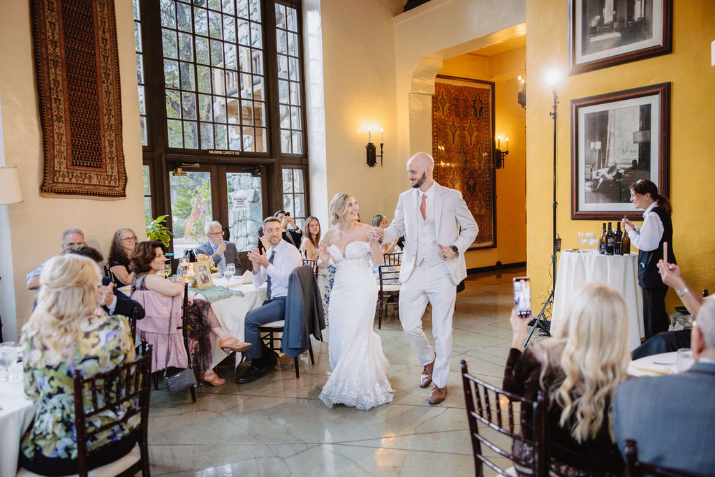 A bride and groom holding hands walk into a brightly lit reception venue. Guests sit around tables, some taking photos, inside a room with large windows and framed pictures on the walls at Ahwahnee Hotel 