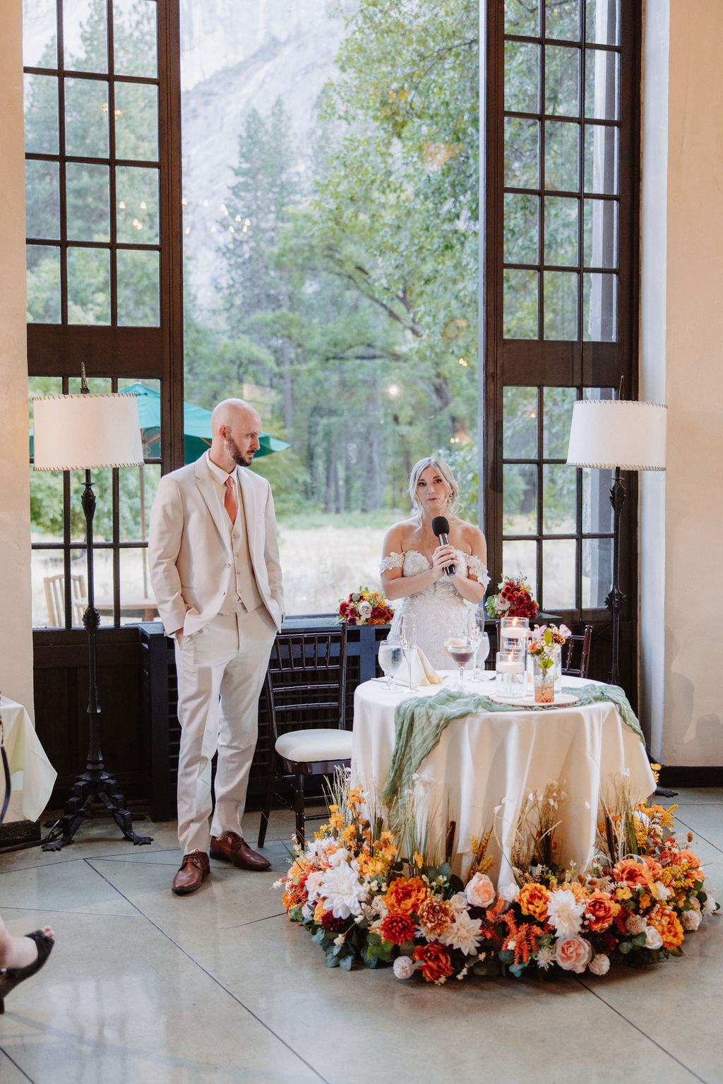 A couple stands near a decorated table with flowers in a bright room, large windows behind them, and trees visible outside.