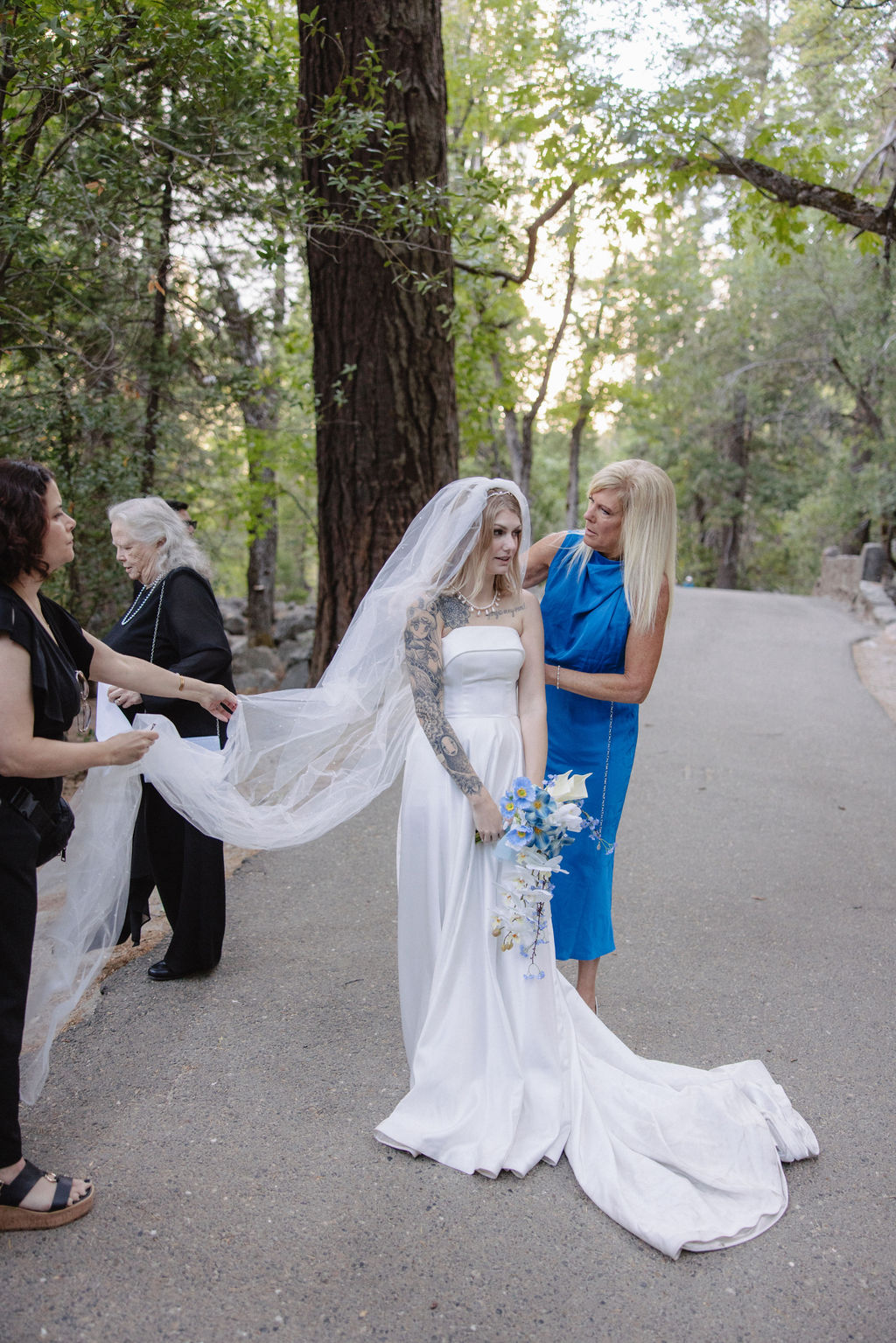 A bride in a white dress stands on a path with two women arranging her veil. She holds a bouquet of blue and white flowers. Trees are in the background.