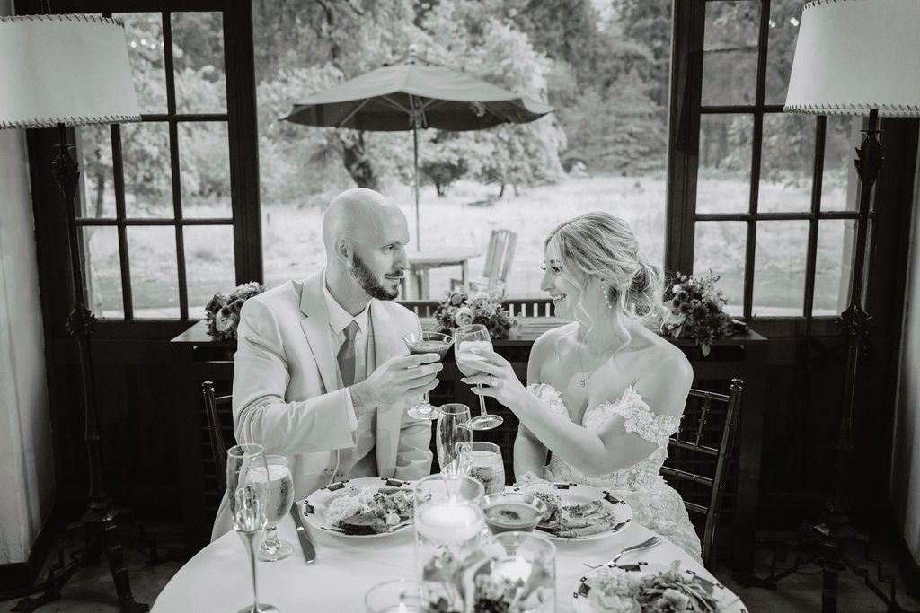 A couple toasting at a table inside near a window, with plates of food and champagne glasses in front of them.