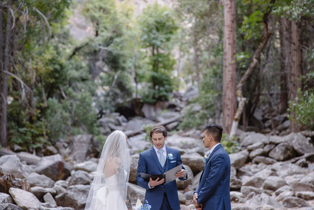 A wedding ceremony taking place outdoors near a waterfall, surrounded by trees. The couple stands with an officiant, while two other people stand nearby.