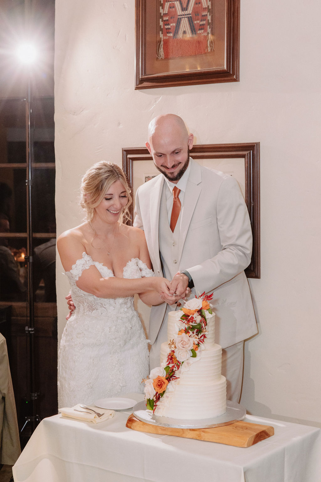 A bride and groom in wedding attire cut a tiered cake decorated with colorful flowers, in a room with framed art on the walls.