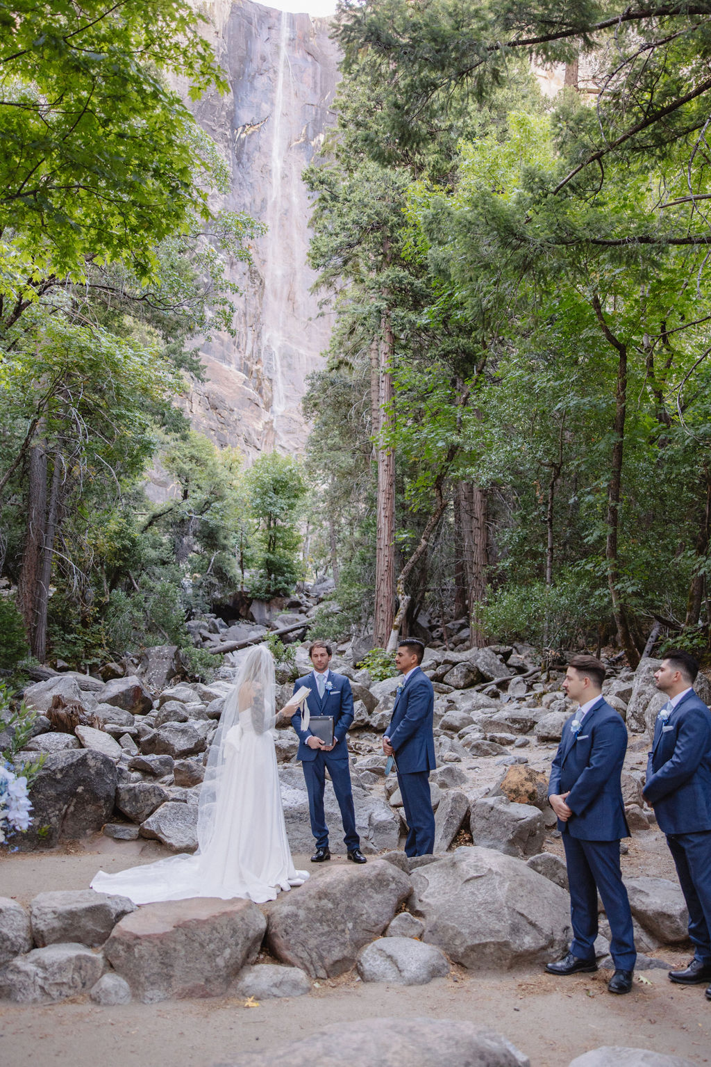 A wedding ceremony taking place outdoors near a waterfall, surrounded by trees. The couple stands with an officiant, while two other people stand nearby.