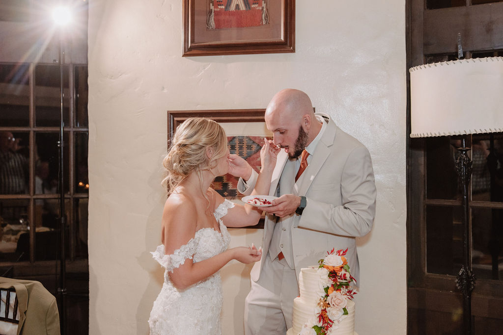 A bride and groom in wedding attire cut a tiered cake decorated with colorful flowers, in a room with framed art on the walls.