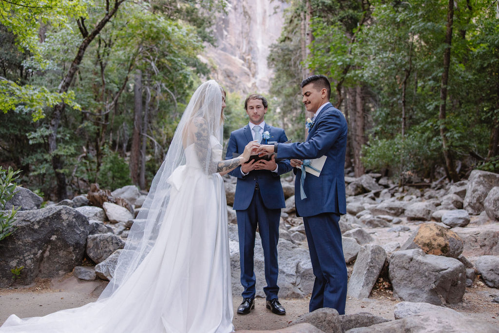 A wedding ceremony taking place outdoors near a waterfall, surrounded by trees. The couple stands with an officiant, while two other people stand nearby.