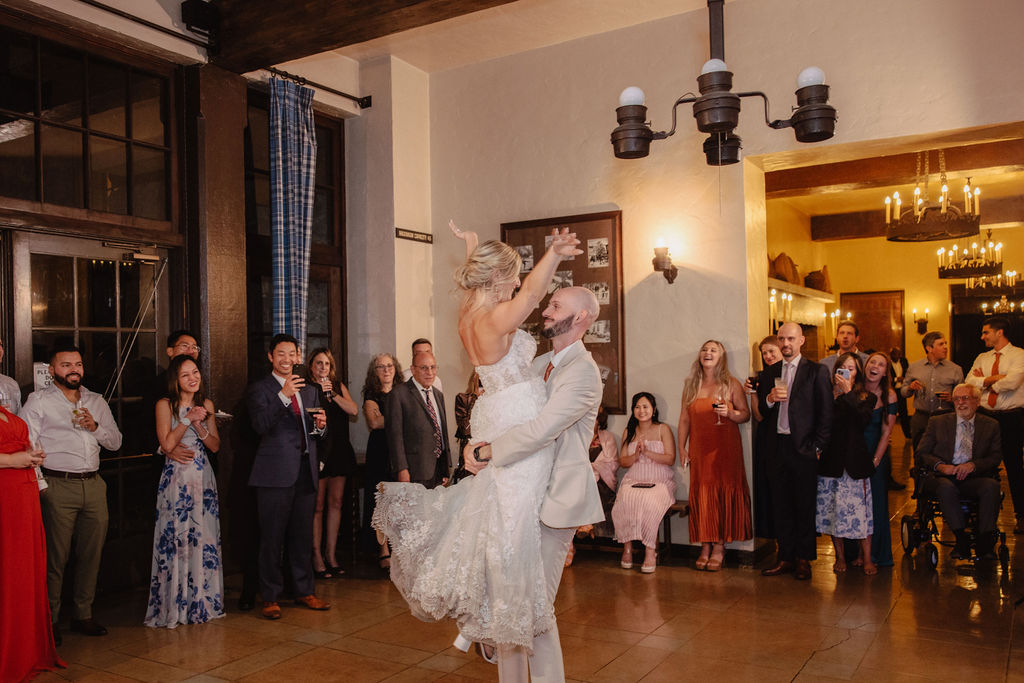 A bride and groom dance joyfully in the center of a room, surrounded by clapping guests.