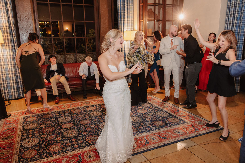 A bride and groom dance joyfully in the center of a room, surrounded by clapping guests.