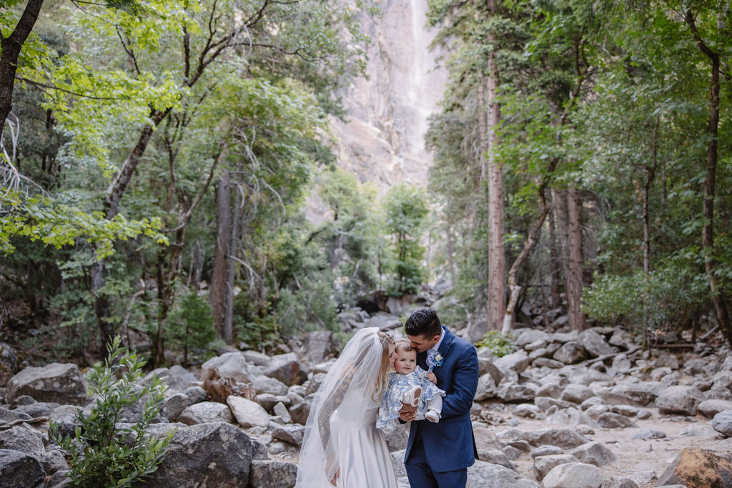 A wedding ceremony taking place outdoors near a waterfall, surrounded by trees. The couple stands with an officiant, while two other people stand nearby.