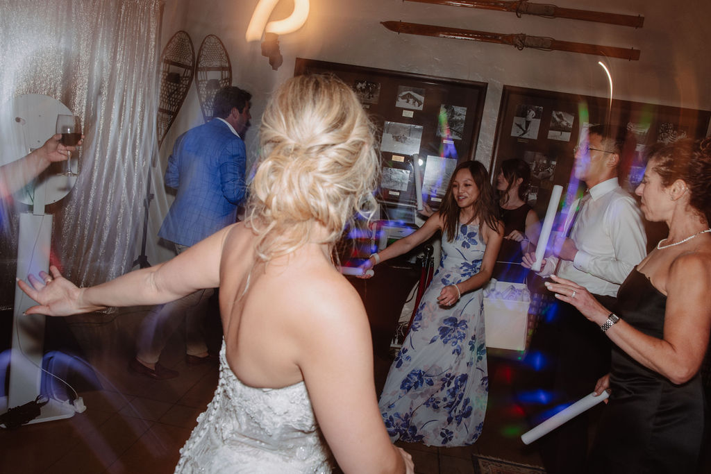 A group of people in formal attire dance at an indoor event with dim lighting. A woman in a white dress is in the foreground.