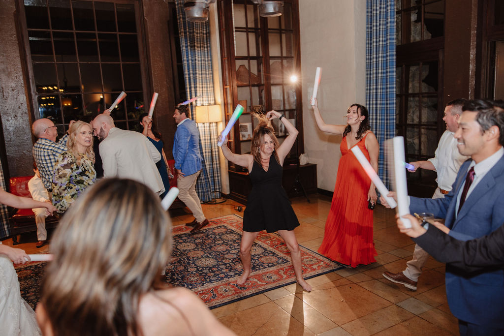 A group of people in formal attire dance at an indoor event with dim lighting. A woman in a white dress is in the foreground.