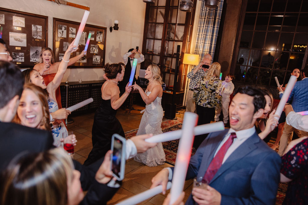 A group of people in formal attire dance at an indoor event with dim lighting. A woman in a white dress is in the foreground.