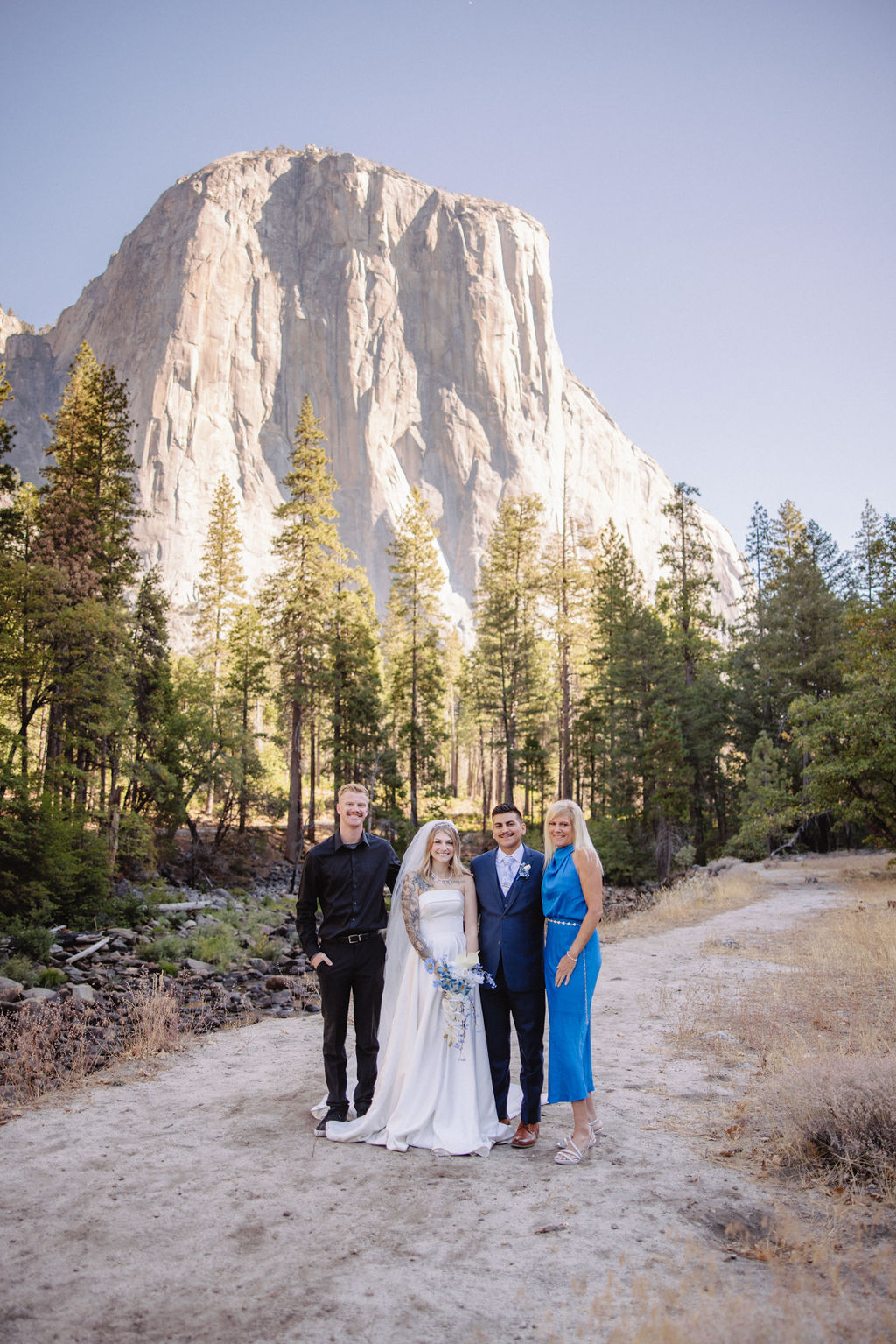 A wedding group poses outdoors, with a bride in white, groom in blue, and two guests. Trees and a large rock formation are in the background under a clear sky.