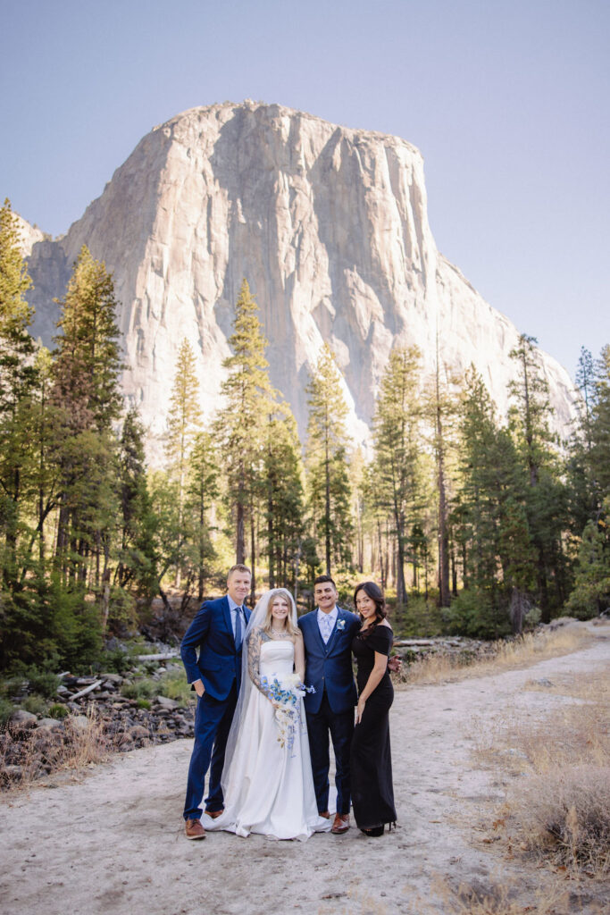 A wedding group poses outdoors, with a bride in white, groom in blue, and two guests. Trees and a large rock formation are in the background under a clear sky.